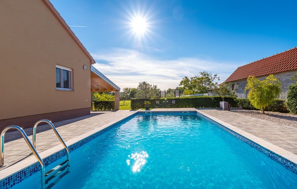 A heated swimming pool is framed by a spacious patio, featuring clear blue water reflecting the sun above. Lush greenery surrounds the area, providing privacy. The house is seen in the background, with a simple facade and large windows inviting natural light.