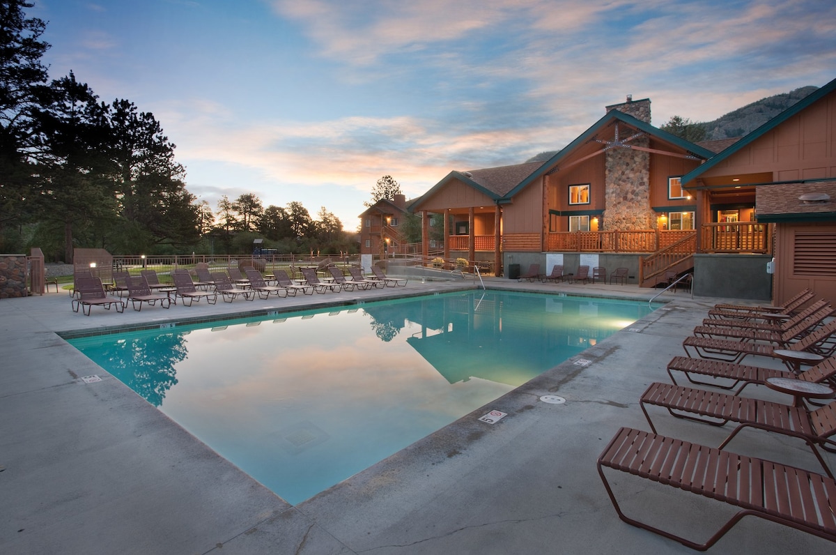 An outdoor heated pool is surrounded by a spacious deck featuring numerous lounge chairs. The pool's clear water reflects the evening sky, while the nearby lodge showcases wooden accents and a welcoming atmosphere. Trees and mountains frame the background, enhancing the serene environment.