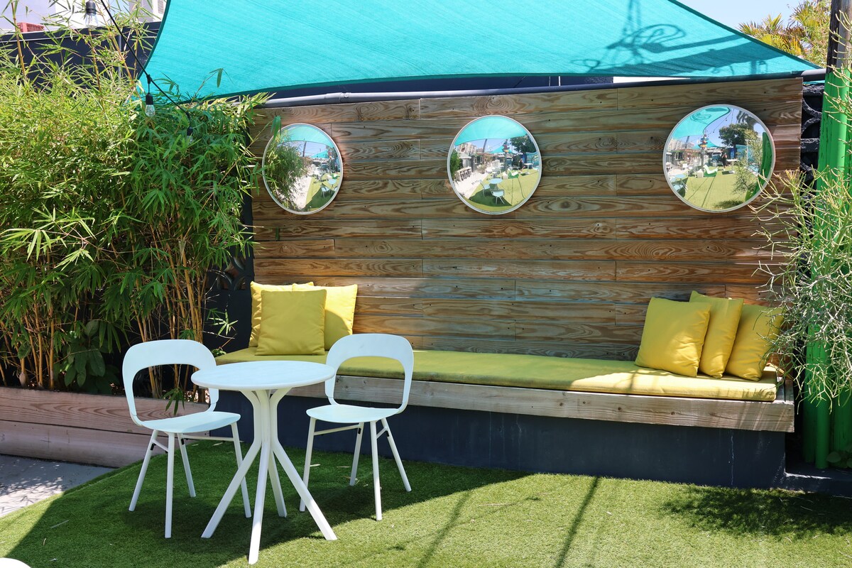 A tranquil garden picnic area features a wooden bench with yellow cushions against a textured wooden wall. Circular mirrors are mounted on the wall, reflecting the surrounding greenery. A small white table and two matching chairs are positioned on the artificial grass beside the bench.