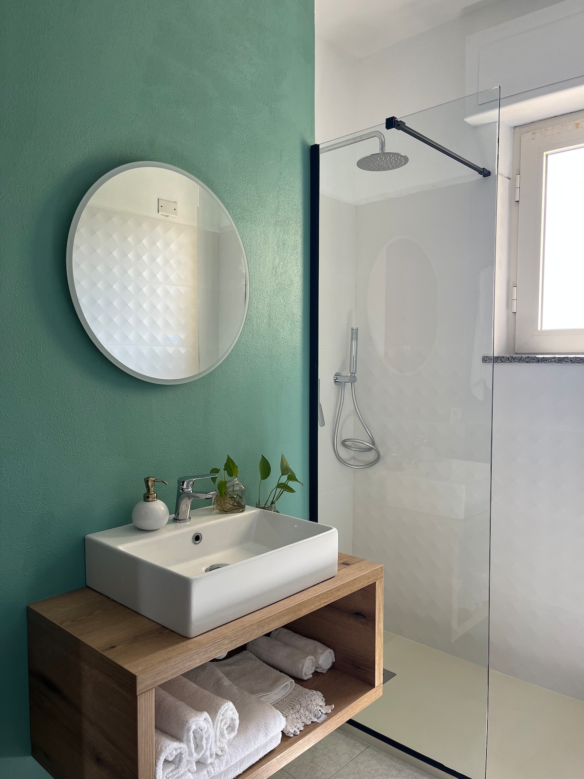 A modern bathroom is showcased with a minimalist design that features a circular mirror above a white vessel sink. Fresh towels are neatly arranged on a wooden shelf below the sink. A walk-in shower with a clear glass enclosure and a handheld showerhead is visible beside a window.