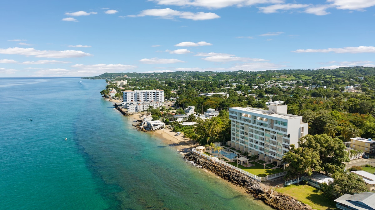 The elevated view captures a beachfront condominium nestled along a serene coastline. Nearby, lush greenery is visible, alongside other structures. The tranquil blue waters stretch toward the horizon, reflecting light under a clear sky dotted with a few clouds.