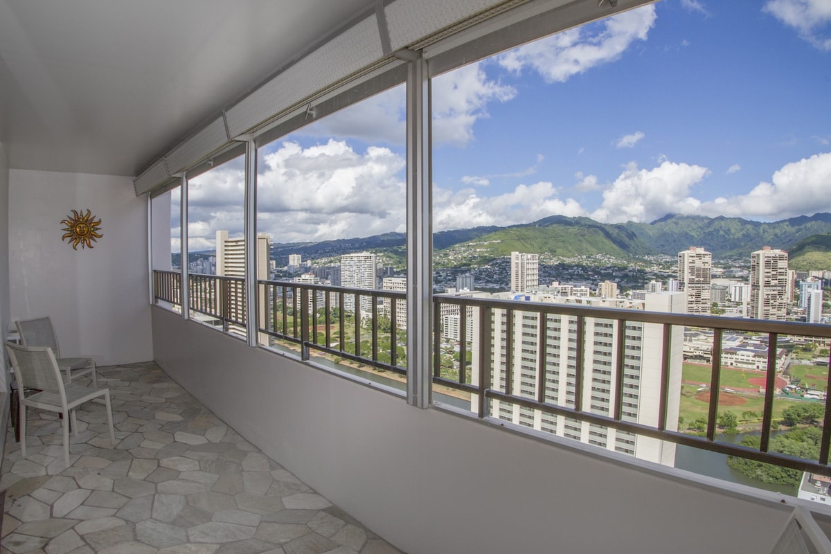 A spacious balcony is presented with a stone floor and railings, offering expansive views of the cityscape and mountains. Large windows allow natural light to fill the area, while a few chairs are positioned for seating. The sky is bright with clouds passing overhead.