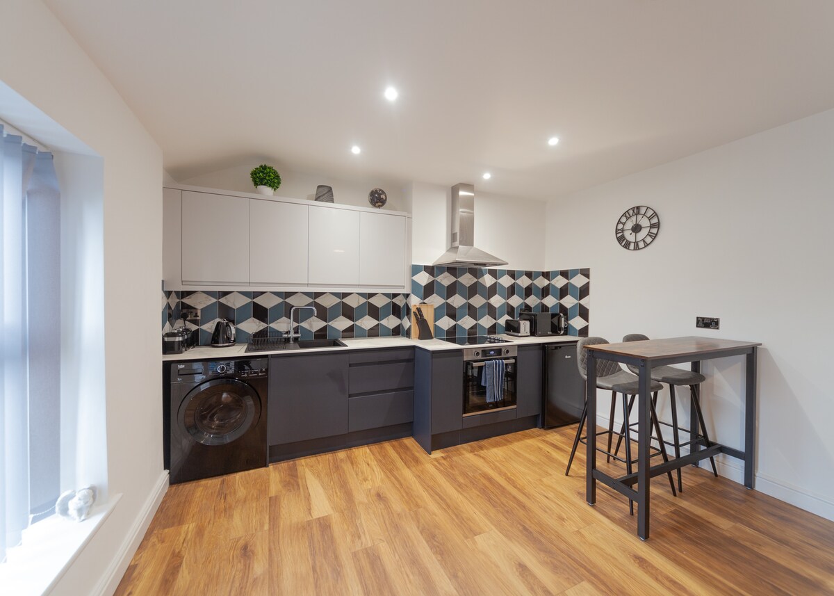 The kitchen features modern cabinetry in neutral tones, highlighted by a geometric backsplash. Equipped with essential appliances, there is a fridge, washer, and oven. A small dining table with two stools is positioned along one side, accompanied by warm wooden flooring that adds to the inviting space.