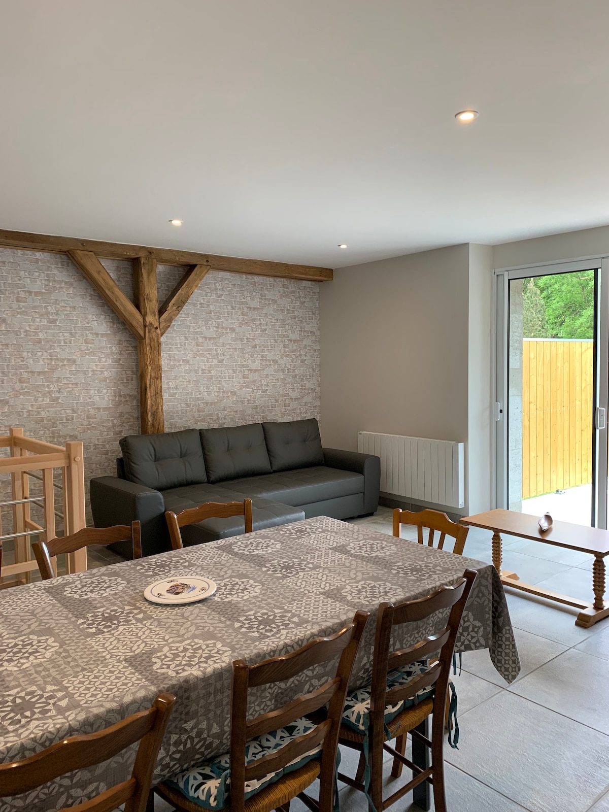A spacious living area is shown, featuring a gray sofa against a textured wall, complemented by wooden beams. A wooden dining table surrounded by chairs is visible, setting a comfortable space for gatherings. Natural light enters through sliding glass doors that lead to an outdoor area.