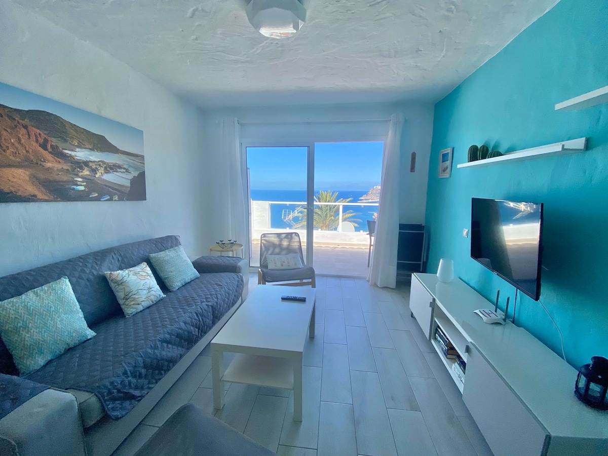 A living area features a comfortable gray sofa with decorative cushions and a light coffee table. Large glass doors provide a view of the sea, allowing natural light to fill the space. A flat-screen TV is positioned against a white entertainment unit.