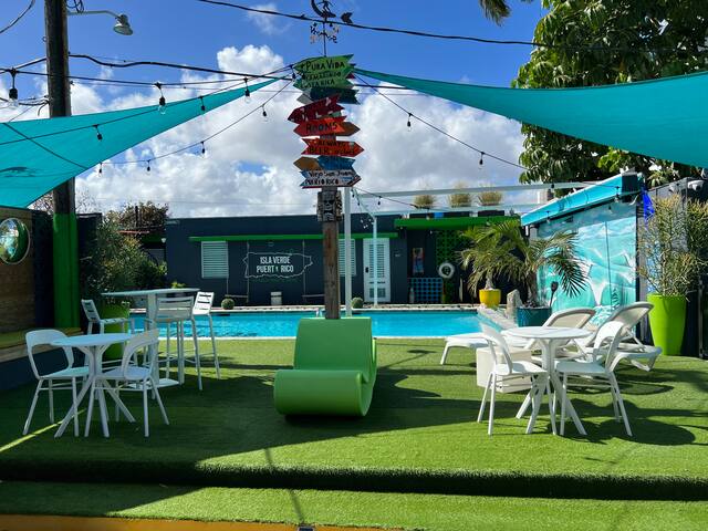 A vibrant outdoor area is seen, featuring colorful lounge chairs and tables placed on green turf. A swimming pool reflects the clear sky, while string lights hang overhead, enhancing the lively atmosphere. Signposts with various destination names adorn the space.
