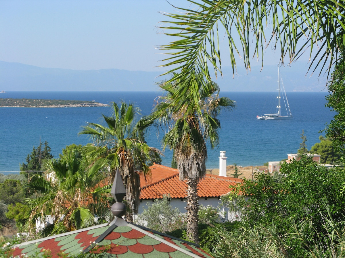 A coastal view is presented, featuring palm trees swaying gently in the breeze. The blue sea is visible, with a sailboat gently anchored offshore and a small island in the distance. A red-tiled rooftop adds a colorful element to the scenery.
