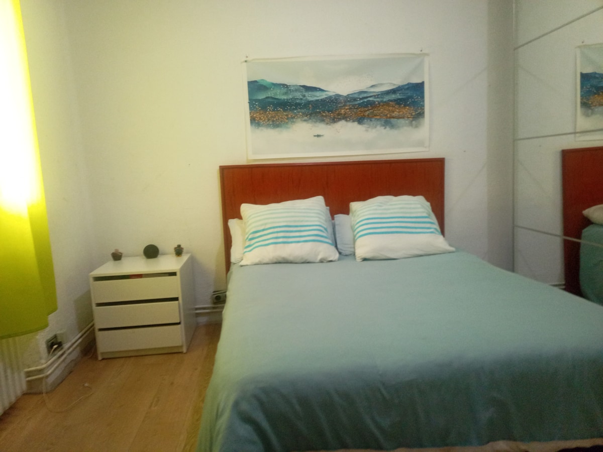 A sleeping area is showcased with a spacious bed featuring light blue bedding and striped pillows. A small bedside table holds decorative items, and a piece of art depicting a landscape is mounted on the wall. Natural light enters through curtains in soft green.