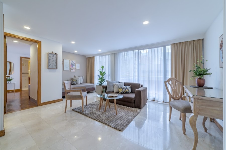 The spacious living area features a comfortable seating arrangement with a sofa and chairs, complemented by a decorative coffee table. Large windows allow natural light to fill the space, while neutral-toned curtains add an element of warmth. A small desk is positioned against the wall.