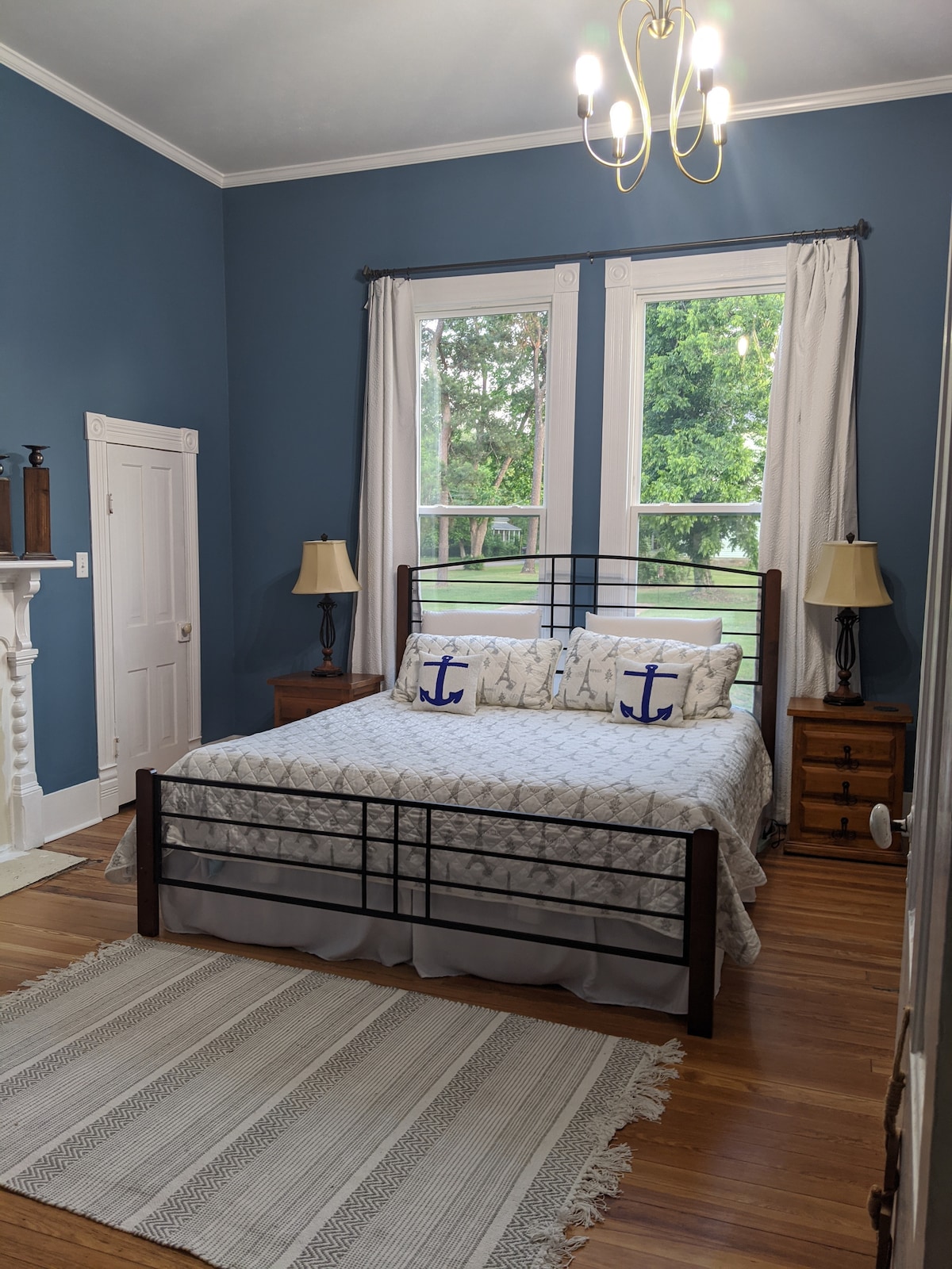 A spacious bedroom features a king bed with decorative pillows, flanked by two bedside tables with lamps. Light blue walls and large windows offer views of the outside greenery, while a light area rug adds comfort underfoot.