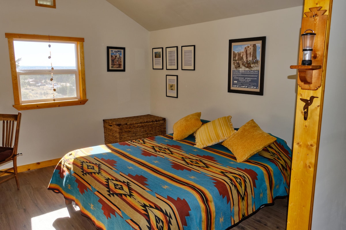 A colorful quilted bedspread adorns a spacious queen bed, complemented by two yellow pillows. A wooden chair and a storage basket are visible, while framed artwork and photographs decorate the walls. A window provides natural light, showcasing scenic views beyond.