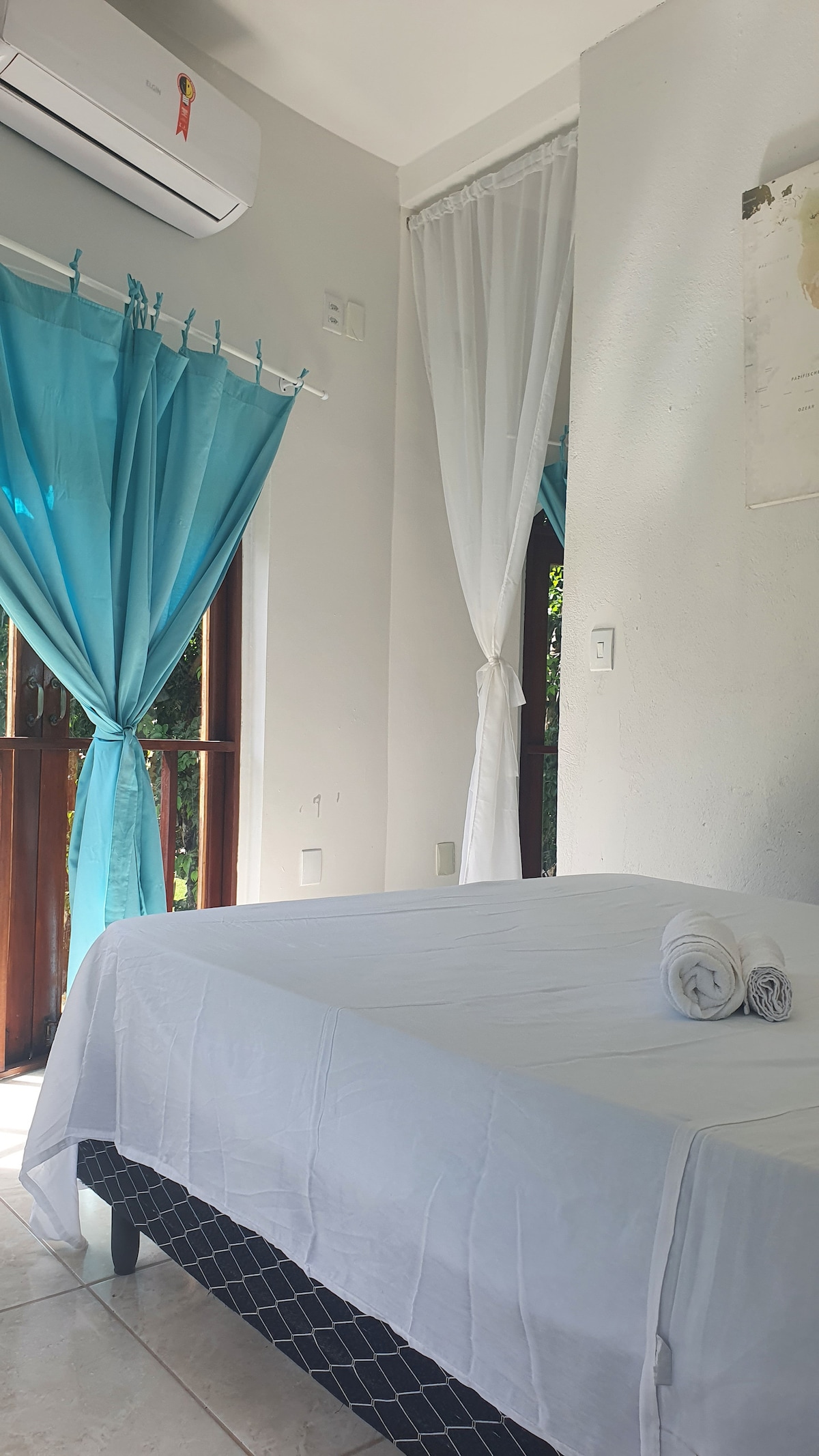 A bright sleeping area features a spacious bed with crisp white linens. Light blue curtains frame the window, allowing natural light to fill the room. A neatly arranged set of towels is placed at the foot of the bed, enhancing the room's simplicity.