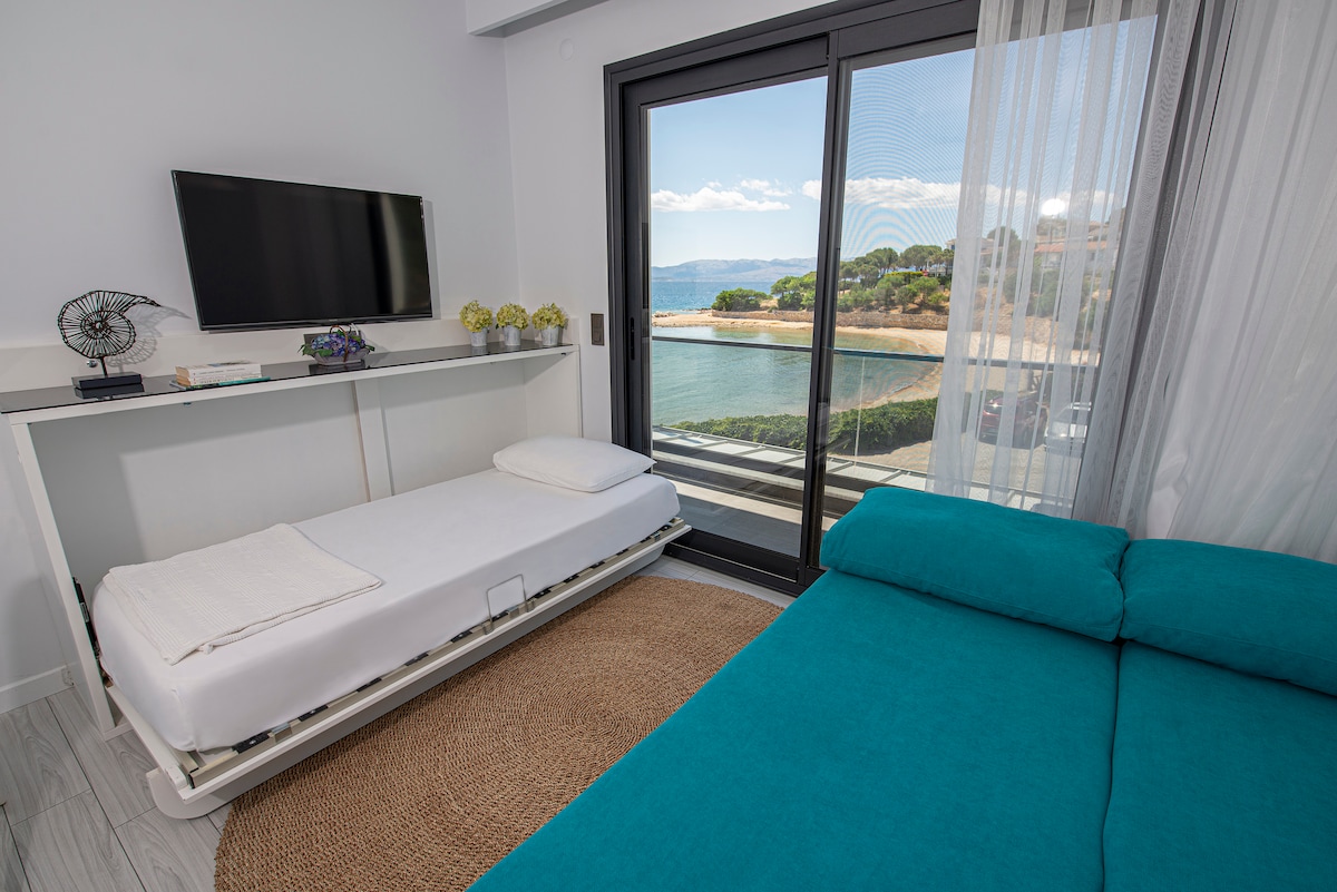 A bright sleeping area is equipped with a spacious blue sofa bed and a white folding bed. A large glass door provides scenic views of the landscape and water. A flat-screen TV is mounted on the wall, with a small shelf displaying decorative items.