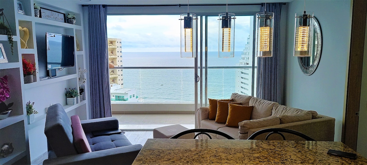 A living area is presented with a panoramic view of the sea through large glass doors. A comfortable light-colored sofa with decorative pillows and a stylish gray chair are arranged around a coffee table. Slim pendant lights hang from the ceiling above a granite kitchen counter.