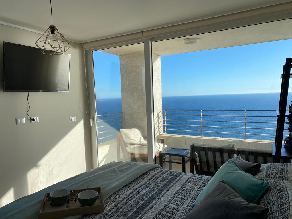 A cozy bedroom features a spacious bed dressed in patterned linens, complemented by decorative cushions. Large glass doors open to a private balcony, offering unobstructed views of the ocean. A wall-mounted TV is visible, enhancing the modern aesthetic of the room.