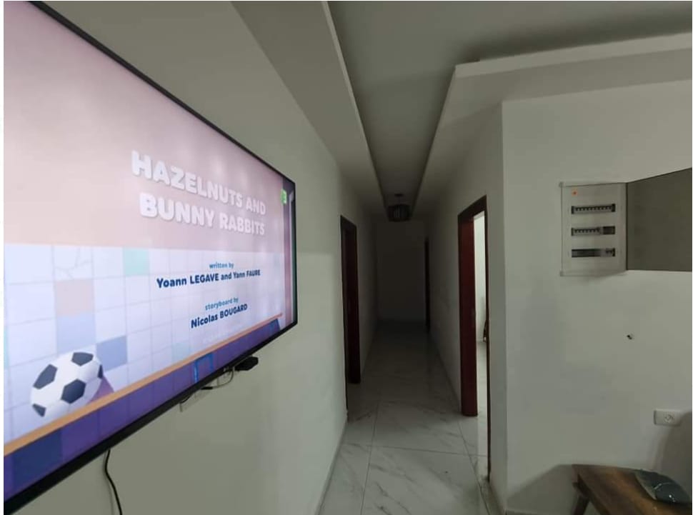 A hallway features a mounted television displaying colorful artwork. The space is illuminated by ceiling lights and has a white marble floor. Doorways lead to rooms on both sides, while a wooden table is positioned on the right side of the image.