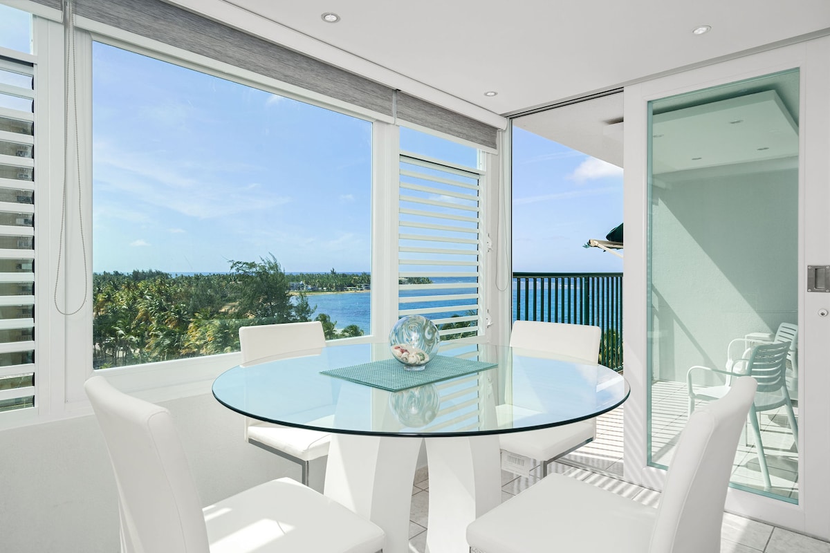 A bright dining area features a round glass table surrounded by four white chairs. Large windows provide expansive views of the ocean and lush greenery. Natural light fills the space, enhancing the modern aesthetic.