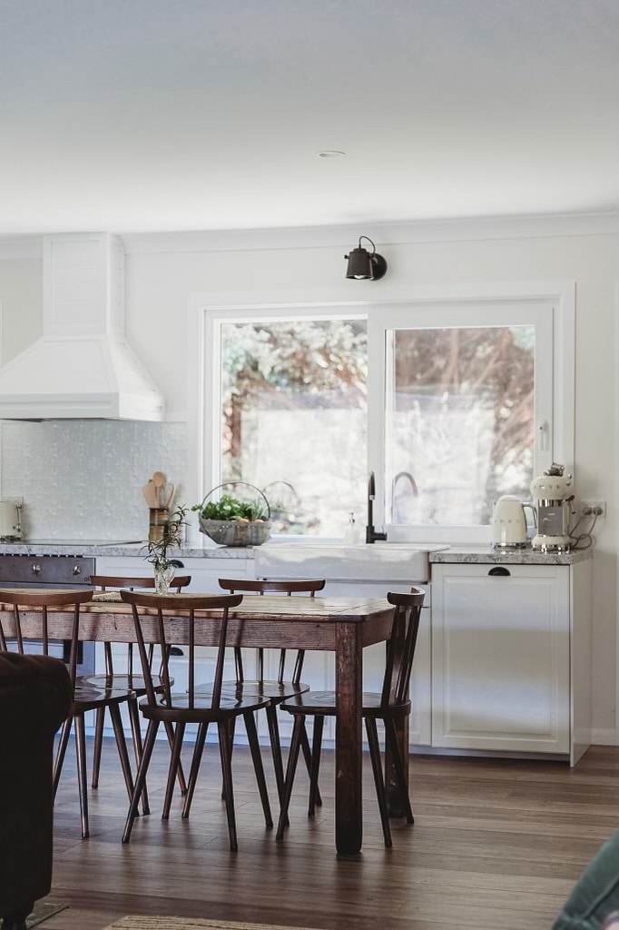 A light-filled kitchen area features a wooden dining table surrounded by six chairs. A window overlooks a garden space, while modern appliances are present on the counter. Neutral tones and natural light create a bright and inviting ambiance.