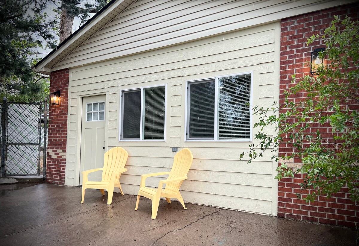 The exterior of the guest house features a welcoming façade with light-colored siding and a brick accent. Two yellow Adirondack chairs are positioned outside, set against a backdrop of large windows that provide natural light into the space.