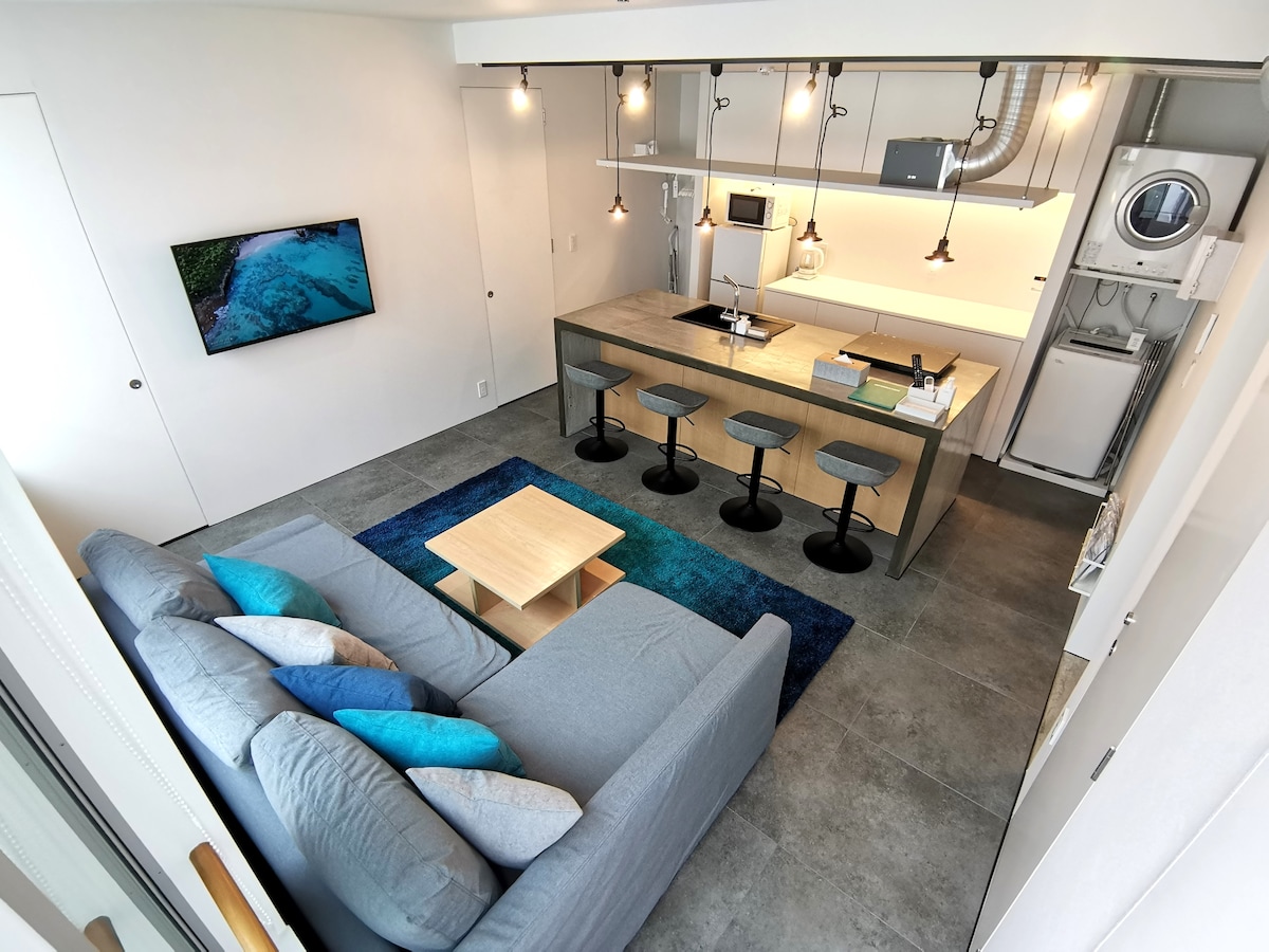 A modern kitchen area features a central island with seating for four. Appliances, including a refrigerator and washing machine, are neatly arranged. A cozy living area with a gray sofa, adorned with several colorful cushions, is positioned adjacent to a wall-mounted TV.