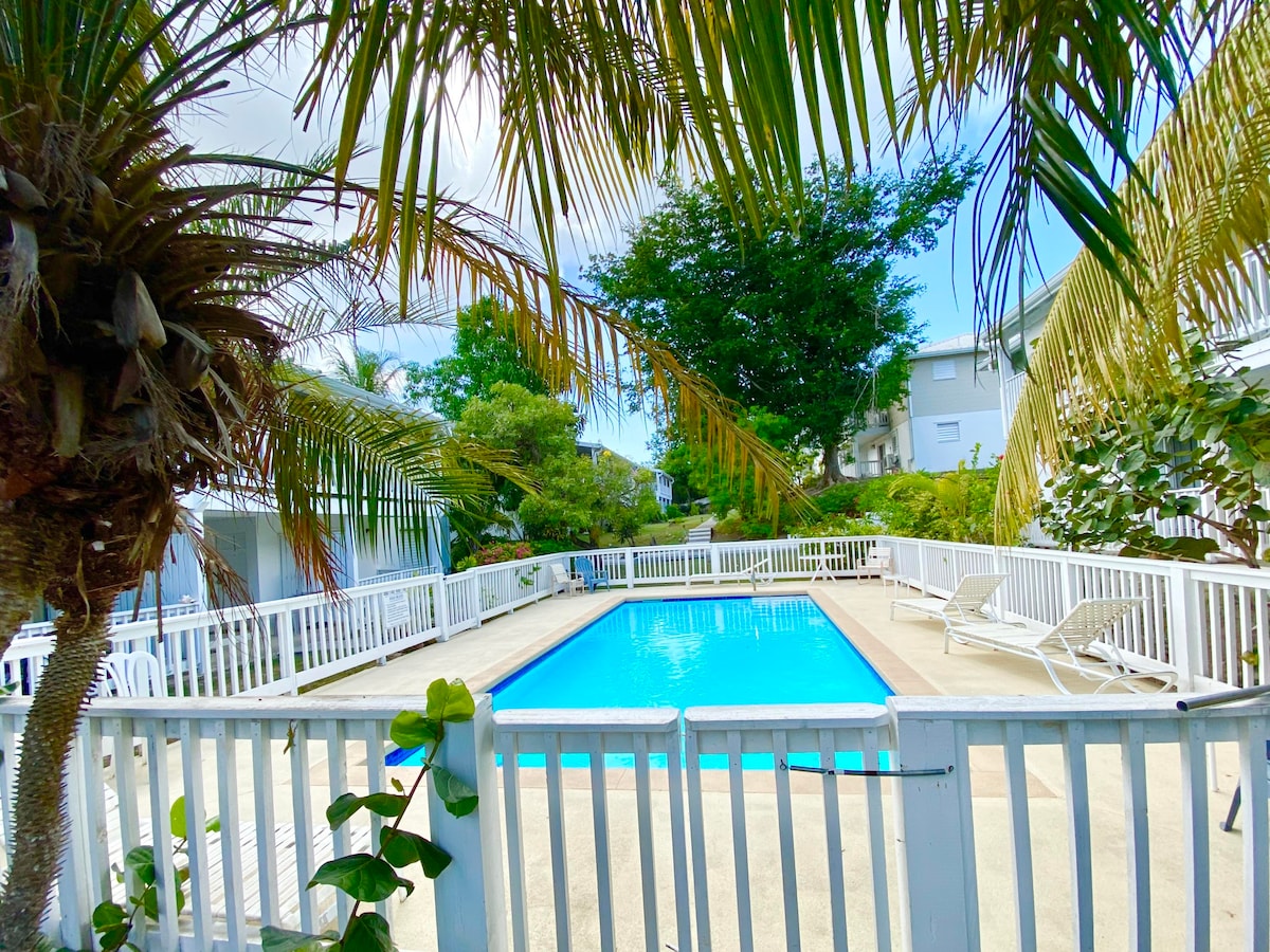 A serene pool is framed by lush palm trees and greenery. The clear blue water reflects the surrounding nature, while lounge chairs are provided around the pool deck. A white fence offers safety and definition to the space, creating a tranquil outdoor area.