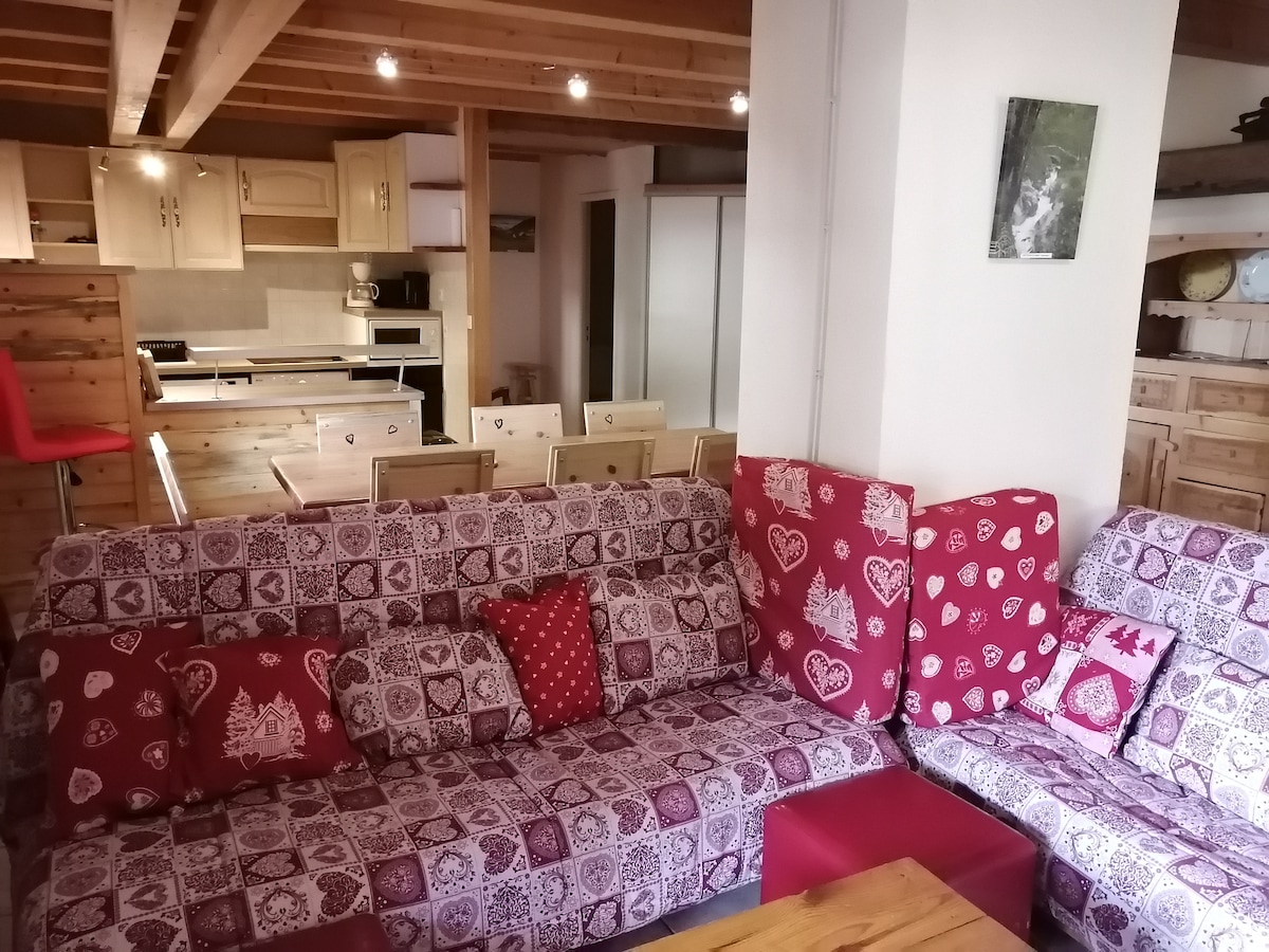 A bright living area is displayed, featuring a spacious sofa adorned with patterned cushions in shades of red and white. Wooden beams line the ceiling, with modern cabinetry visible in the background. Soft lighting enhances the inviting ambiance of the space.