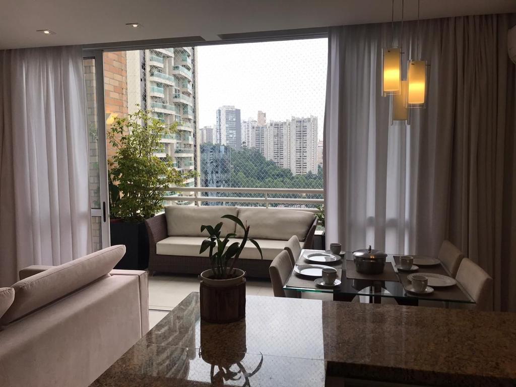 A bright living area is showcased with large glass doors leading to a balcony. Elegant curtains frame the space, complementing a modern sofa and dining table set for four. A potted plant adds a touch of greenery, enhancing the view of the city skyline beyond.