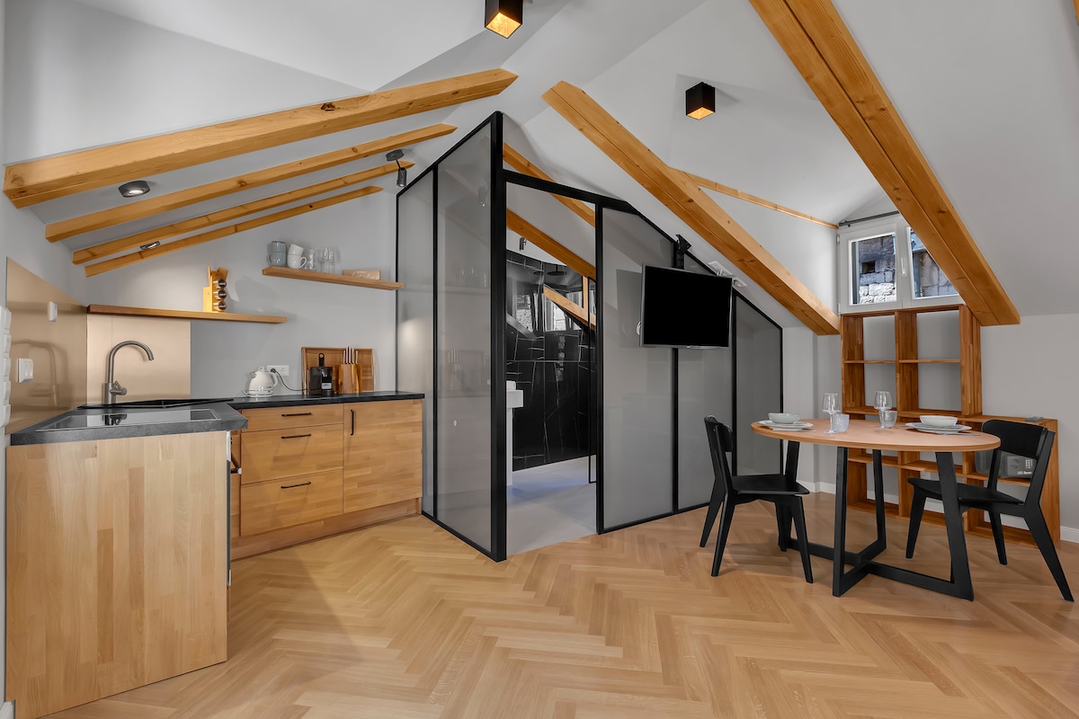 An open-plan living space features wooden beams and modern finishes. A kitchenette with light wood cabinetry is visible, alongside a dining table set for four. A television is mounted on the wall, and a glass partition separates the shower area from the rest of the space.
