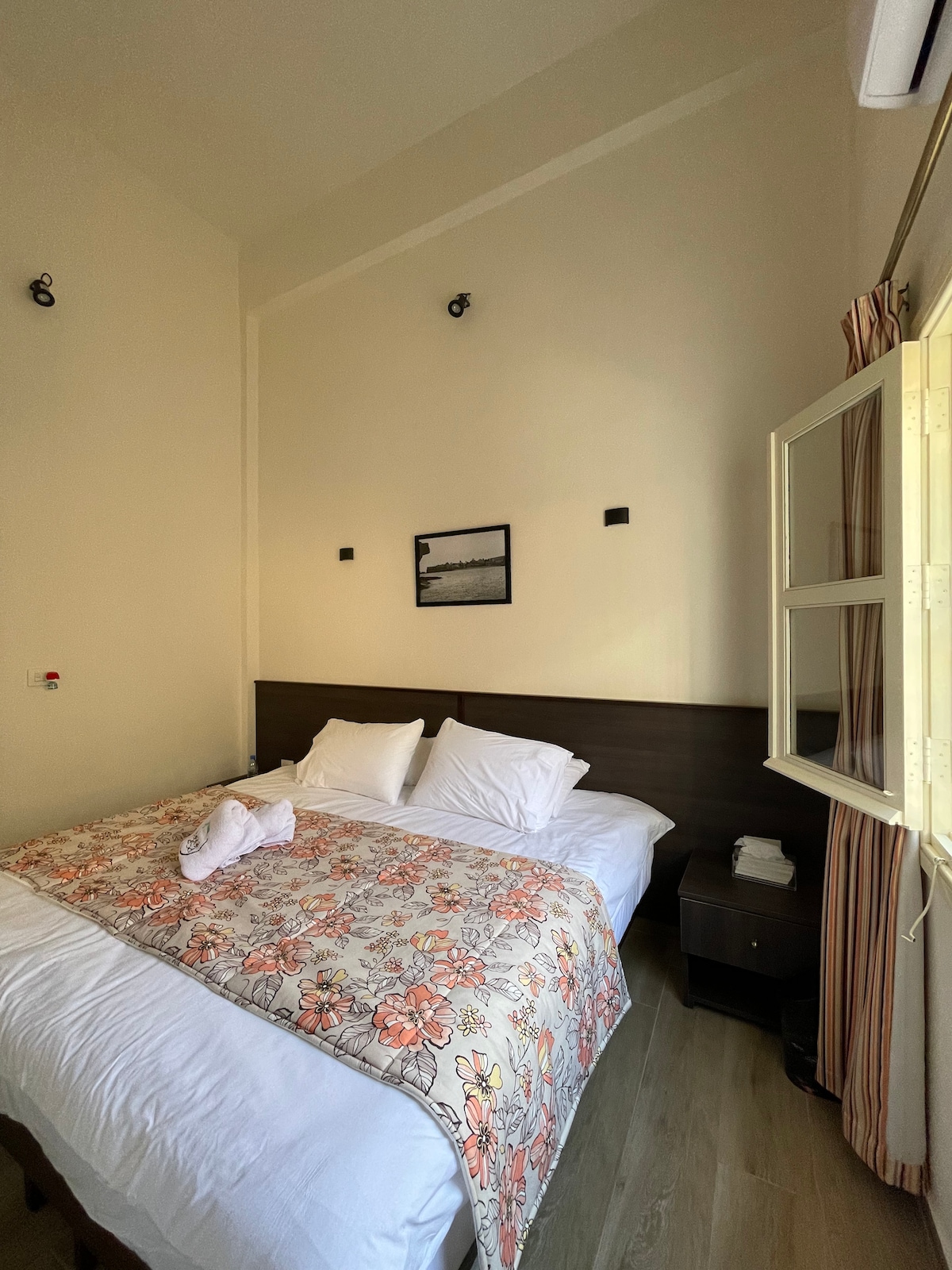A cozy room features a spacious bed layered with a floral blanket and multiple pillows. Light streams through a window with beige curtains, illuminating the modern decor and wood furnishings. A photograph hangs above the headboard, adding a touch of character to the inviting space.