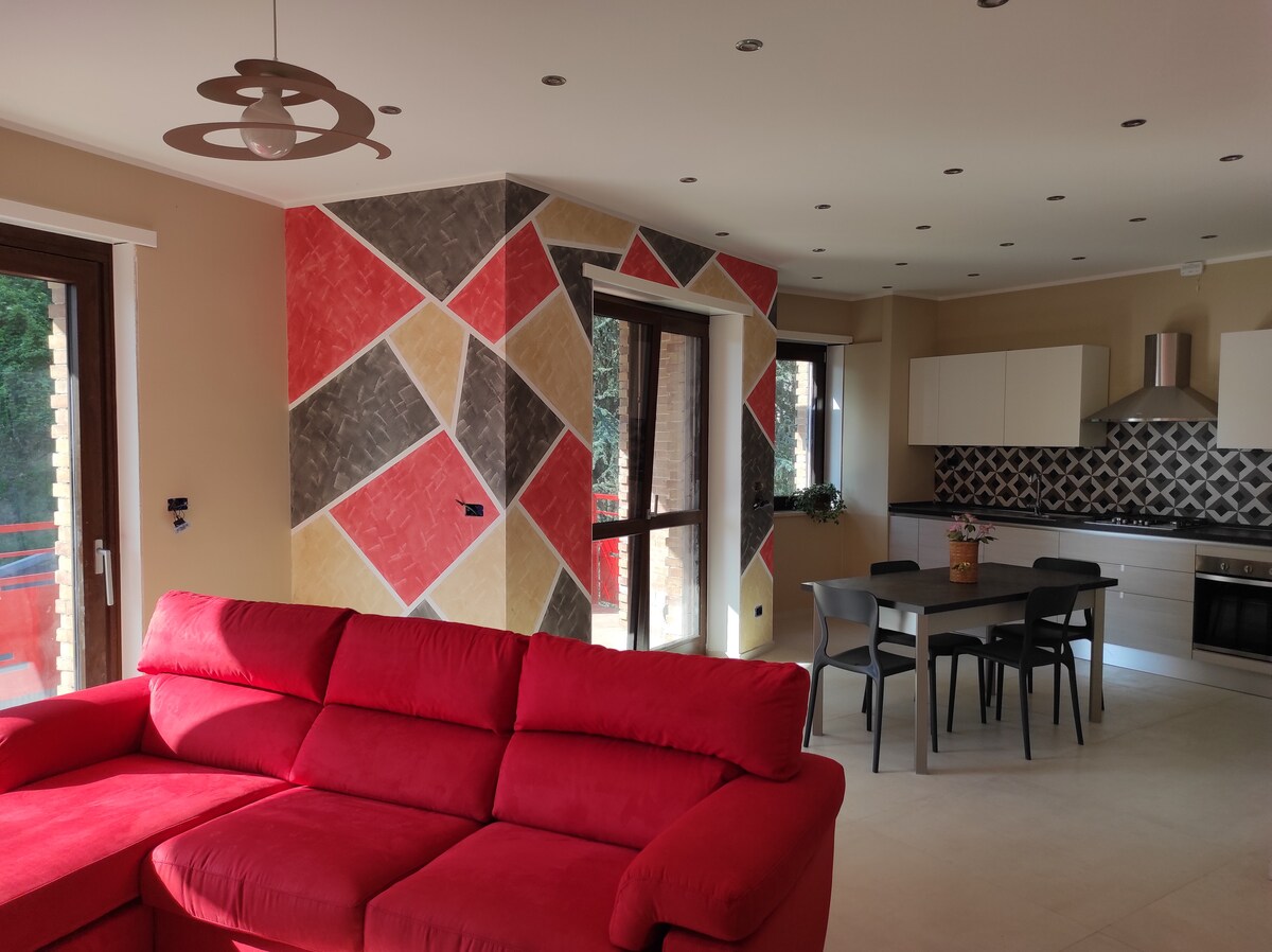 A vibrant living area features a red sectional sofa, providing a bold contrast to the multi-colored accent wall. A round dining table with black chairs is visible, alongside modern kitchen appliances and cabinetry. Large windows allow natural light to illuminate the space.