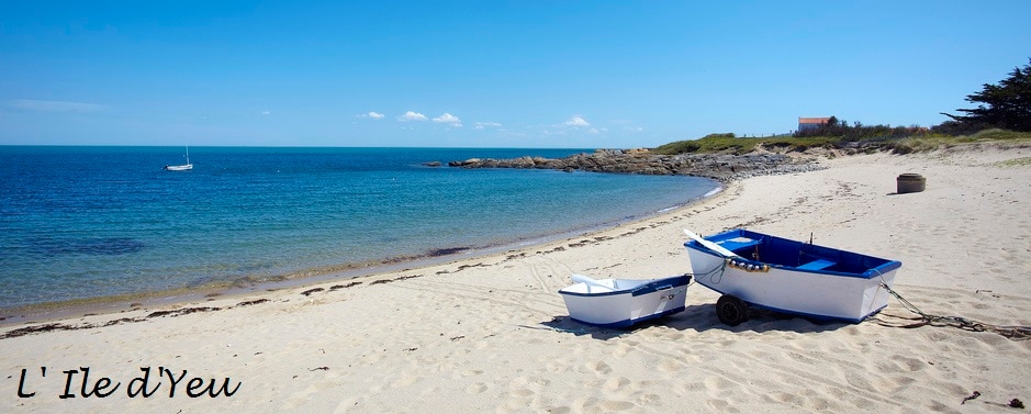 A sandy beach is bordered by calm blue waters under a clear sky. Two small boats are stationed on the shore, with a distant sailboat visible on the horizon. The scenic view includes a rocky coastline and hints of greenery in the background.