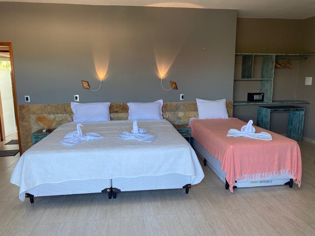 A bright sleeping area is furnished with two spacious beds, one adorned with a light pink blanket and the other with a white coverlet. Natural light enters through the window, and a minimalist shelving unit is present alongside a small microwave.