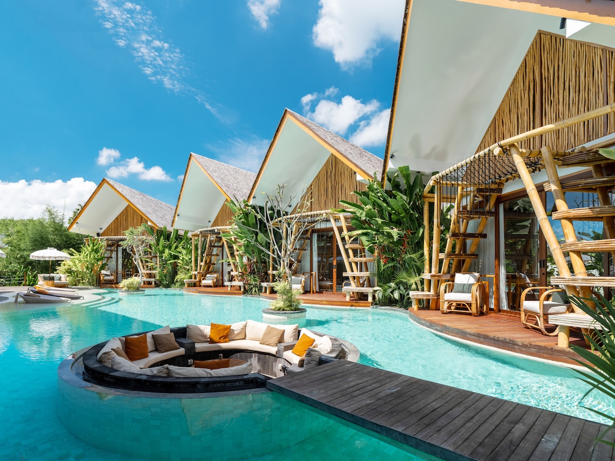 A serene outdoor area showcases eco-friendly bungalows surrounded by lush greenery. A circular lounge area is set within a turquoise pool, featuring a cozy arrangement of cushions. The bright blue sky and light clouds provide a cheerful backdrop to this tranquil setting.