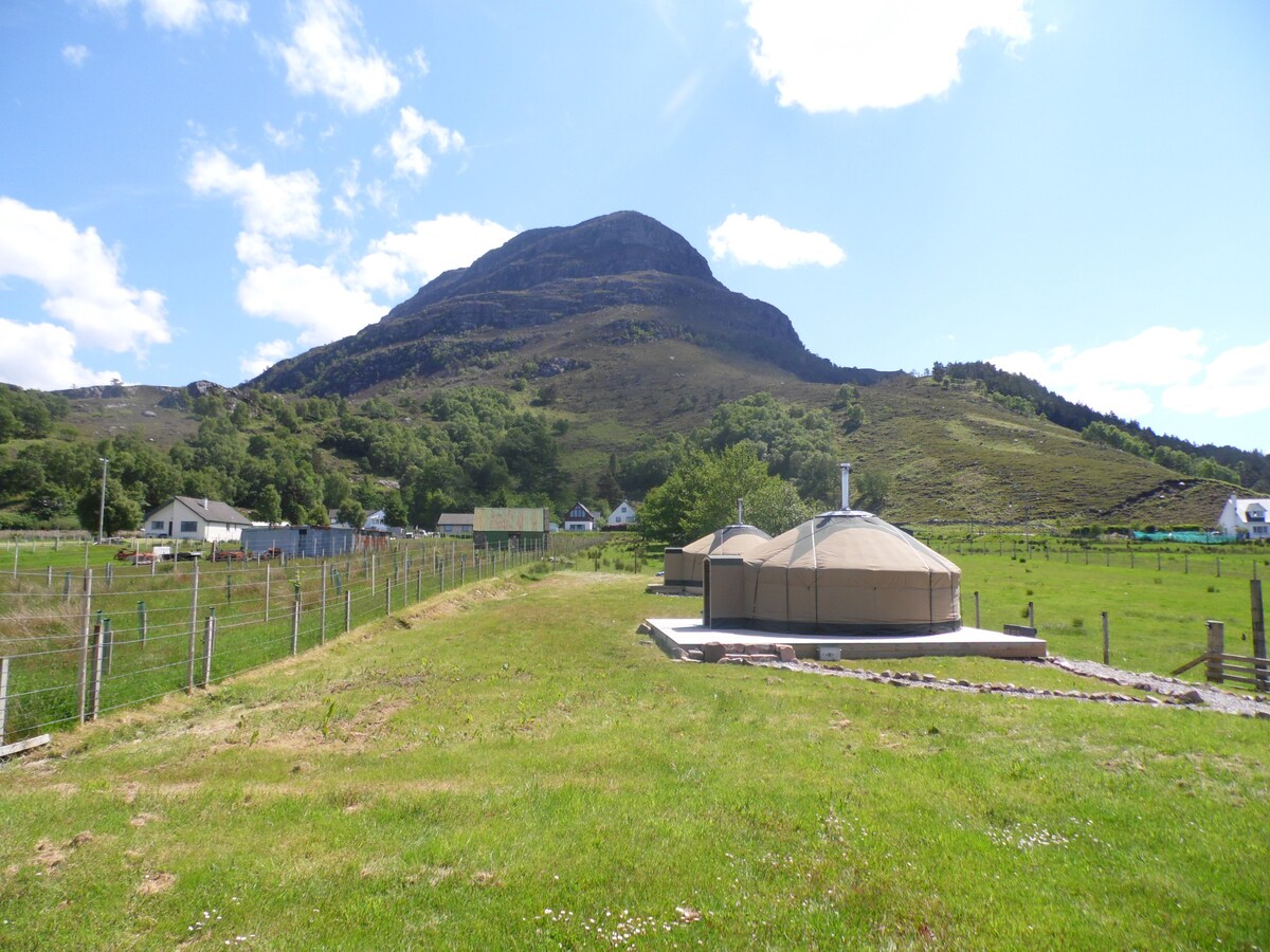 A newly built Mongolian yurt stands on a grassy area against a backdrop of rolling hills. The structure is situated on a family croft, with Beinn Shieldaig prominently rising in the background. Clear blue skies and scattered clouds create a serene atmosphere.