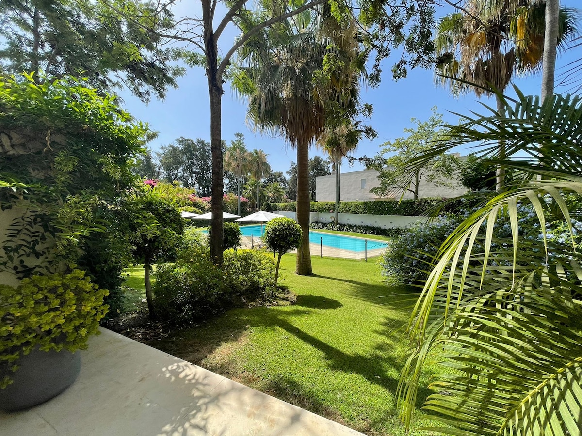 A well-maintained garden area is seen, featuring lush greenery and colorful plants. The outdoor pool is visible, surrounded by lounge chairs and umbrellas, with a tranquil atmosphere enhanced by tall palm trees and a clear blue sky.