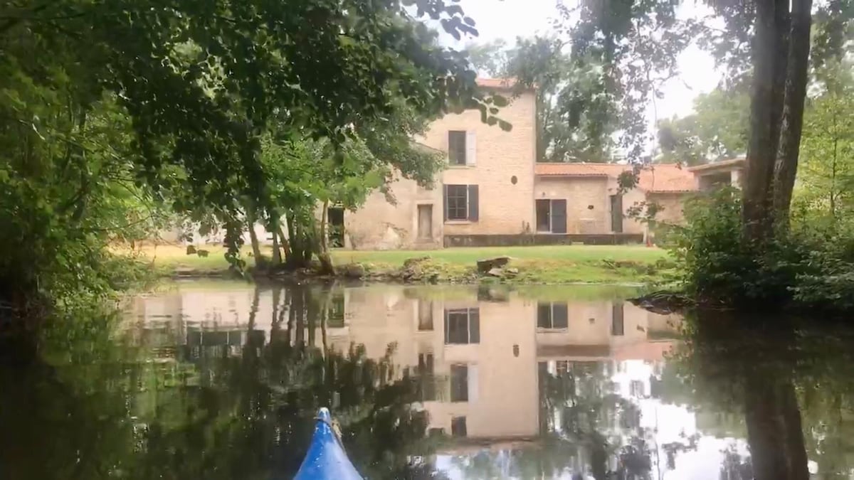 The semi-detached house is seen from across the tranquil water, with its stone facade and large windows reflecting on the surface. Surrounded by greenery, the property features a spacious lawn and is nestled among trees, creating a serene and natural setting.