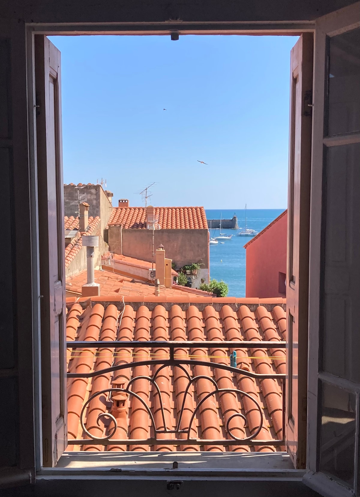A view is framed by open wooden shutters, showcasing a vibrant blue sea and rooftops with red tiles. Boats are visible on the water, and a clear sky is dotted with a few birds, highlighting the charm of the coastal village.