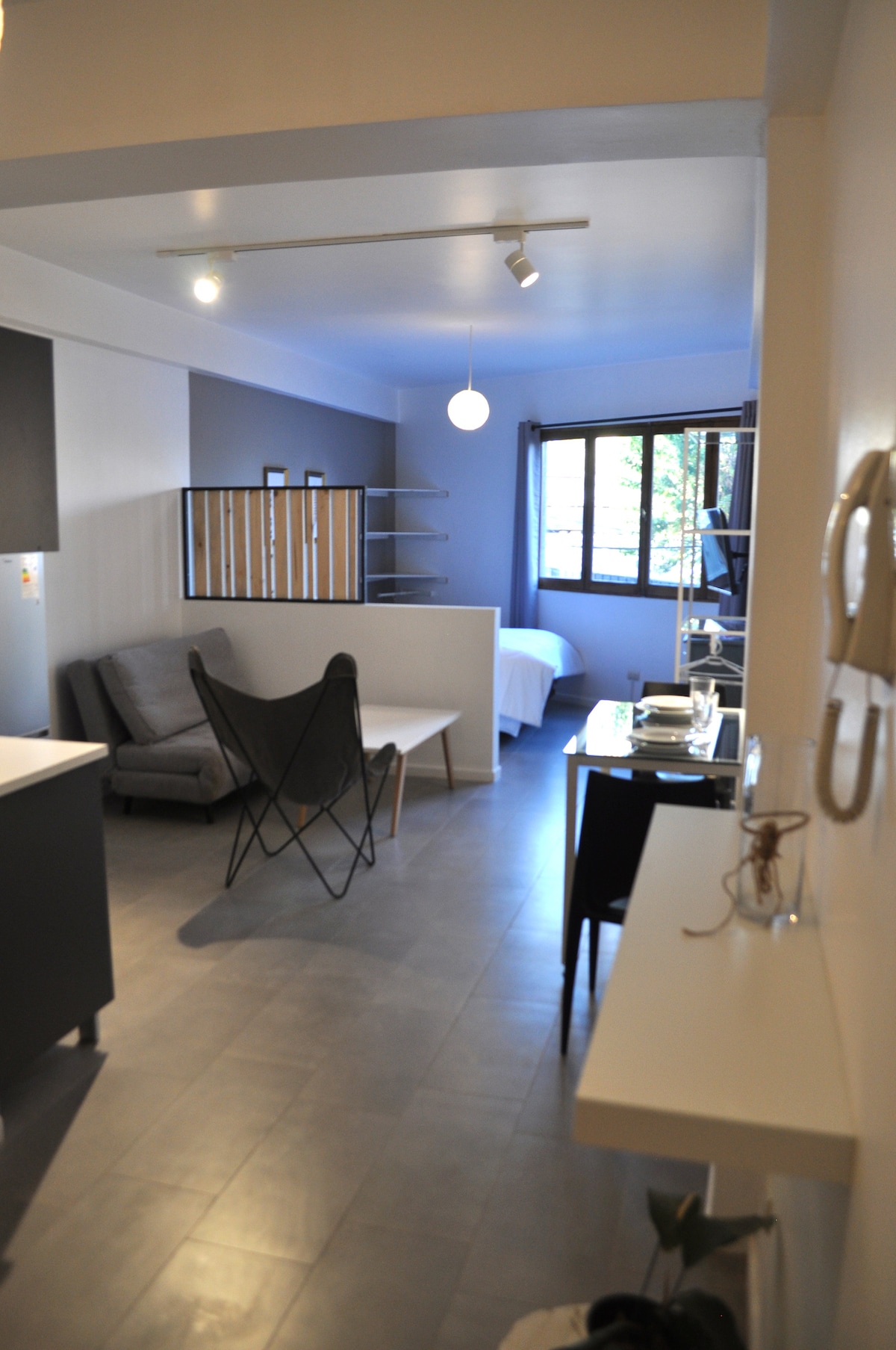 A spacious studio layout is displayed, featuring a soft-gray sofa and a minimalist table in the foreground. The sleeping area can be seen in the background, separated by a decorative partition. Large windows allow natural light to brighten the room, providing views of outdoor greenery.