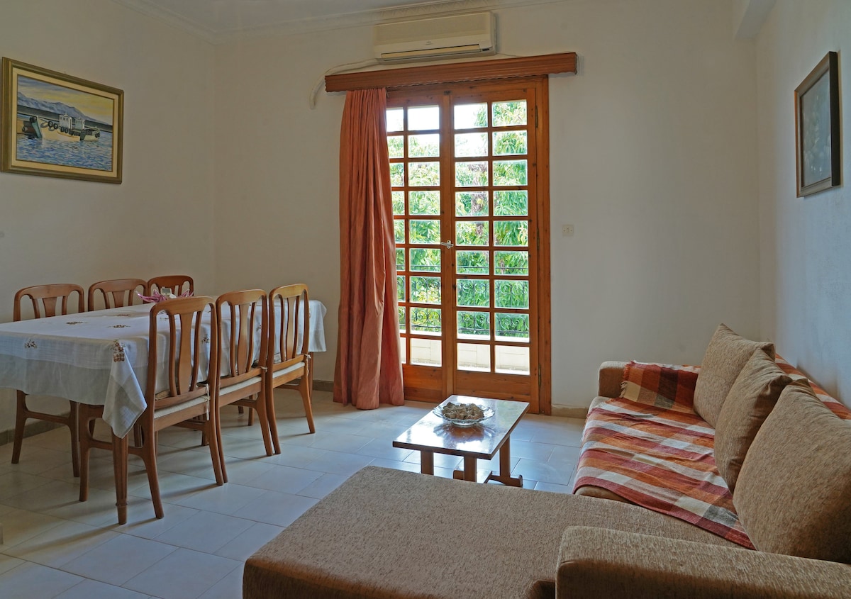 A light-filled living area features a combination of a cozy sofa and a wooden dining table with matching chairs. The sliding glass doors lead to a balcony, allowing for outdoor views. A decorative table holds an arrangement of natural materials.