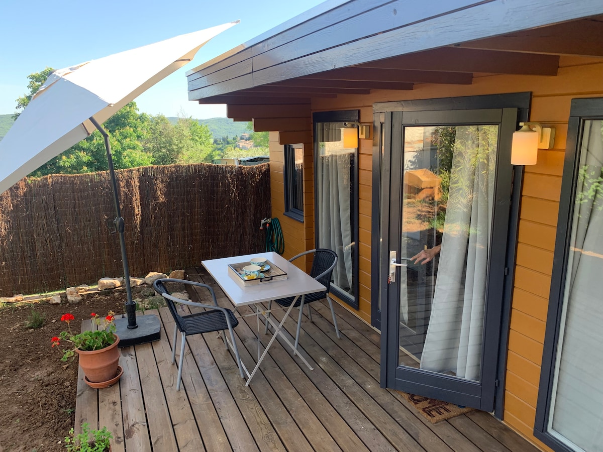 A private terrace features a wooden deck, complemented by a small table set for two and black folding chairs. An umbrella provides shade, while a potted flower adds a touch of greenery. The entrance boasts sliding glass doors, inviting natural light into the interior.