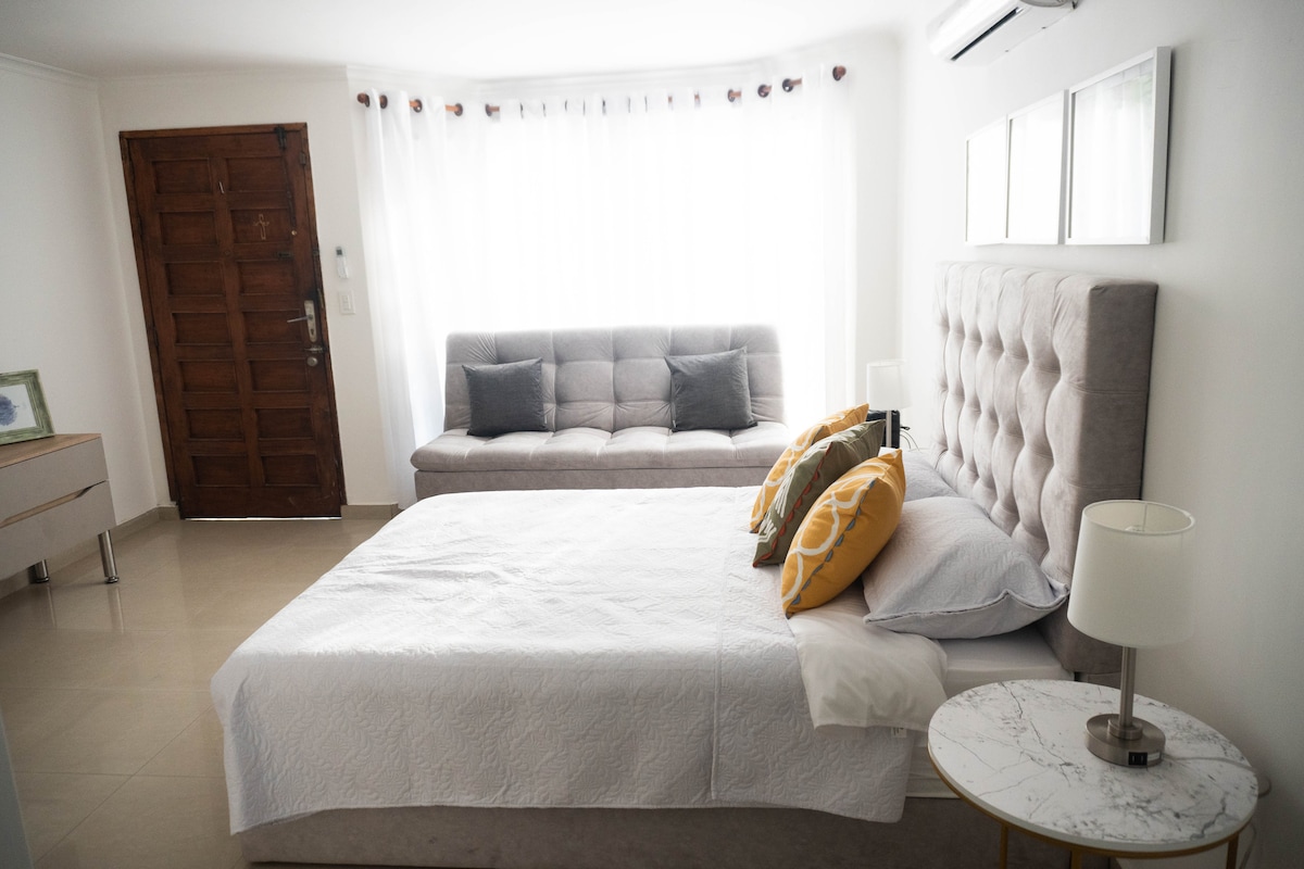 A bright and airy space features a spacious bed covered with a white quilt, complemented by two decorative pillows. A gray sofa adds additional seating, and a small marble-topped table sits next to the bed. Natural light filters through sheer curtains.