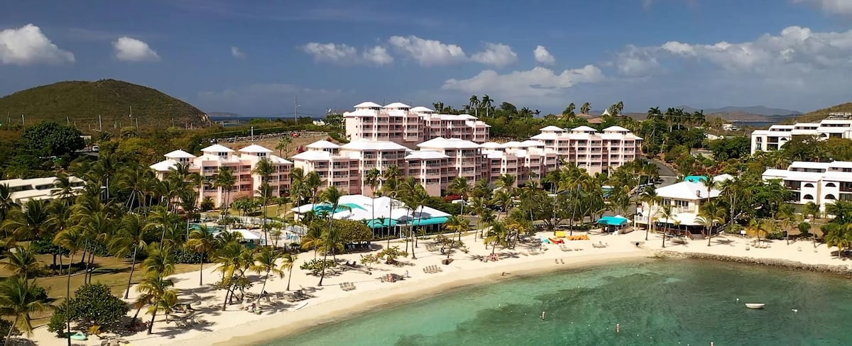 An aerial view captures a resort surrounded by palm trees. The beachfront features a sandy shore leading to clear water. A collection of colorful lounge chairs is positioned near the beachfront, while the resort buildings display a light pink hue against the blue sky.