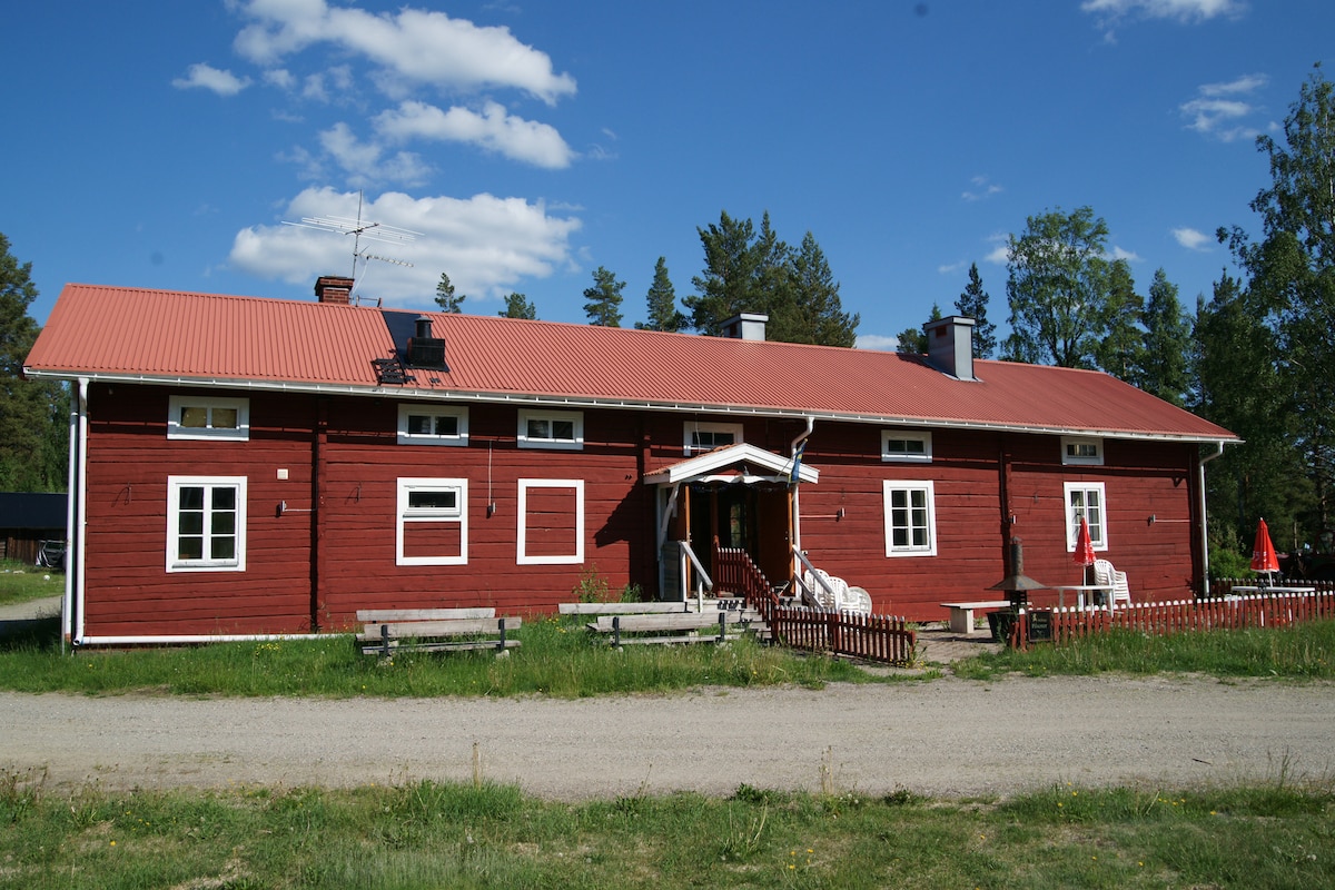 The exterior of a traditional red timber house is showcased, featuring a welcoming porch with steps leading up to the entrance. The building is flanked by several windows and has a sloping roof, set against a backdrop of greenery and a blue sky.