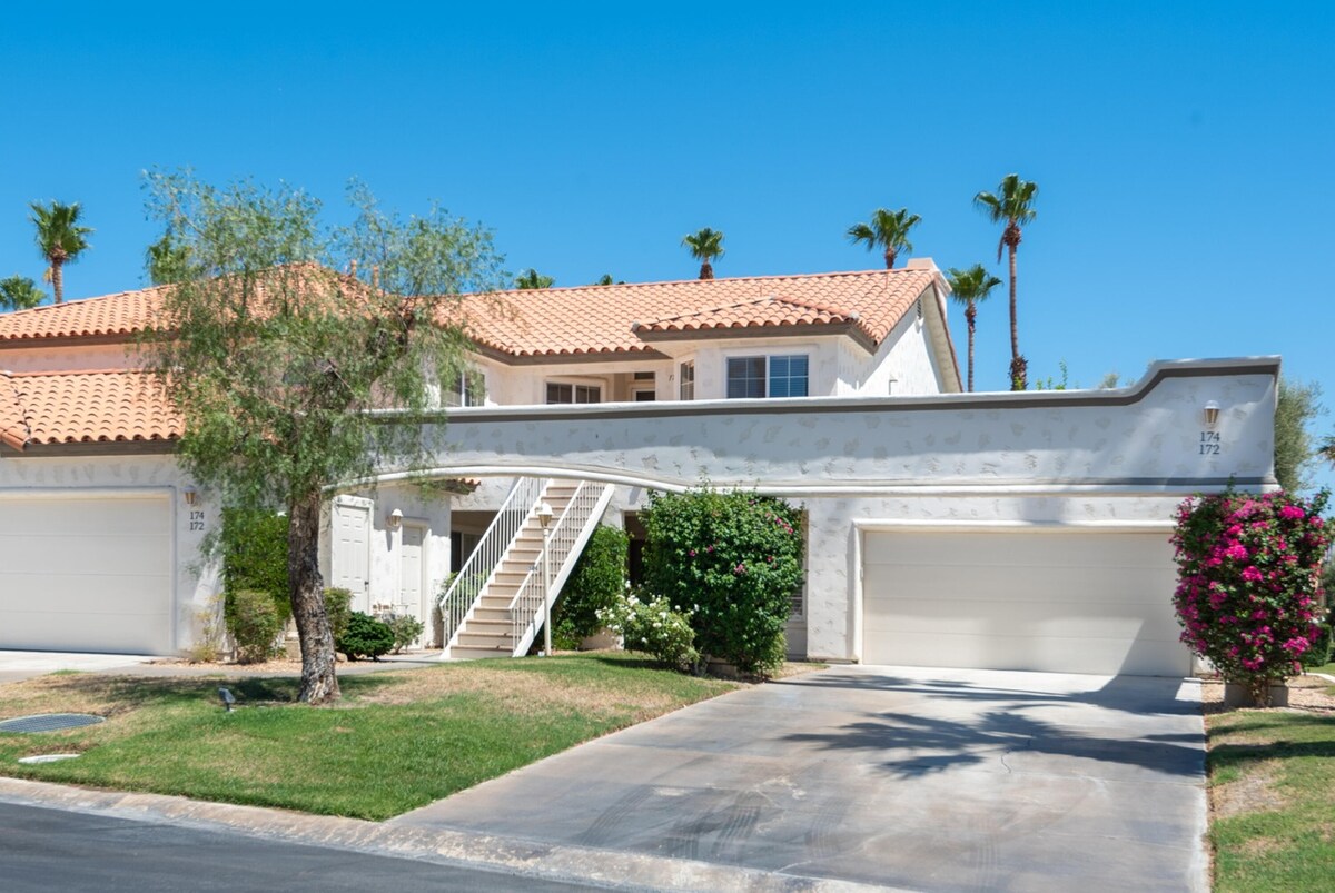A two-story villa is shown with a stucco exterior and a red-tiled roof. The entrance features stairs leading to the upper level, flanked by lush greenery and blooming flowers. Two garage doors are visible, providing ample parking space.
