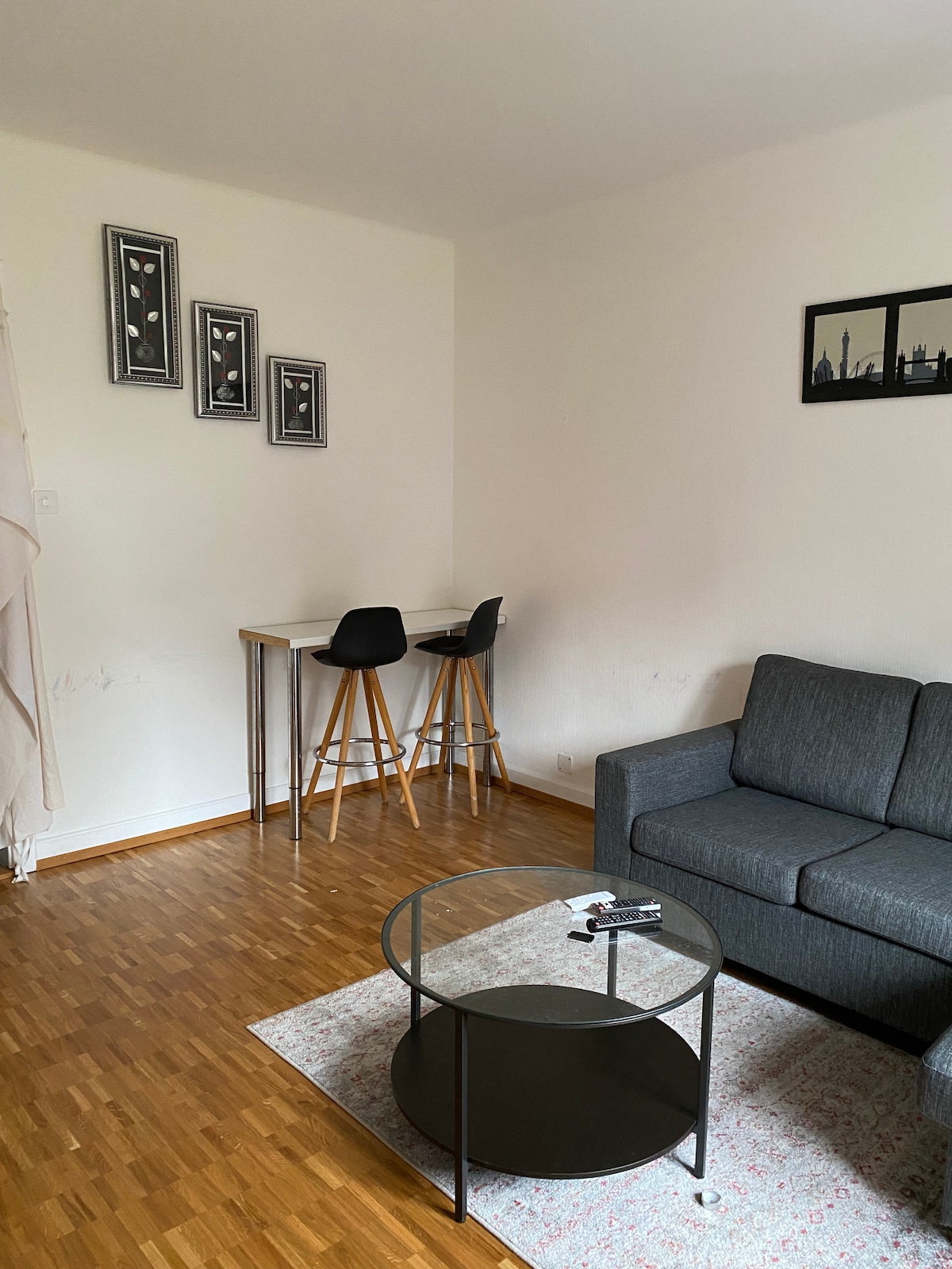 A cozy living area features a gray sofa and a circular black coffee table atop a textured rug. In the background, a small dining setup includes a glass table with two black chairs. Three framed artworks hang above the table, complementing the minimalist decor.