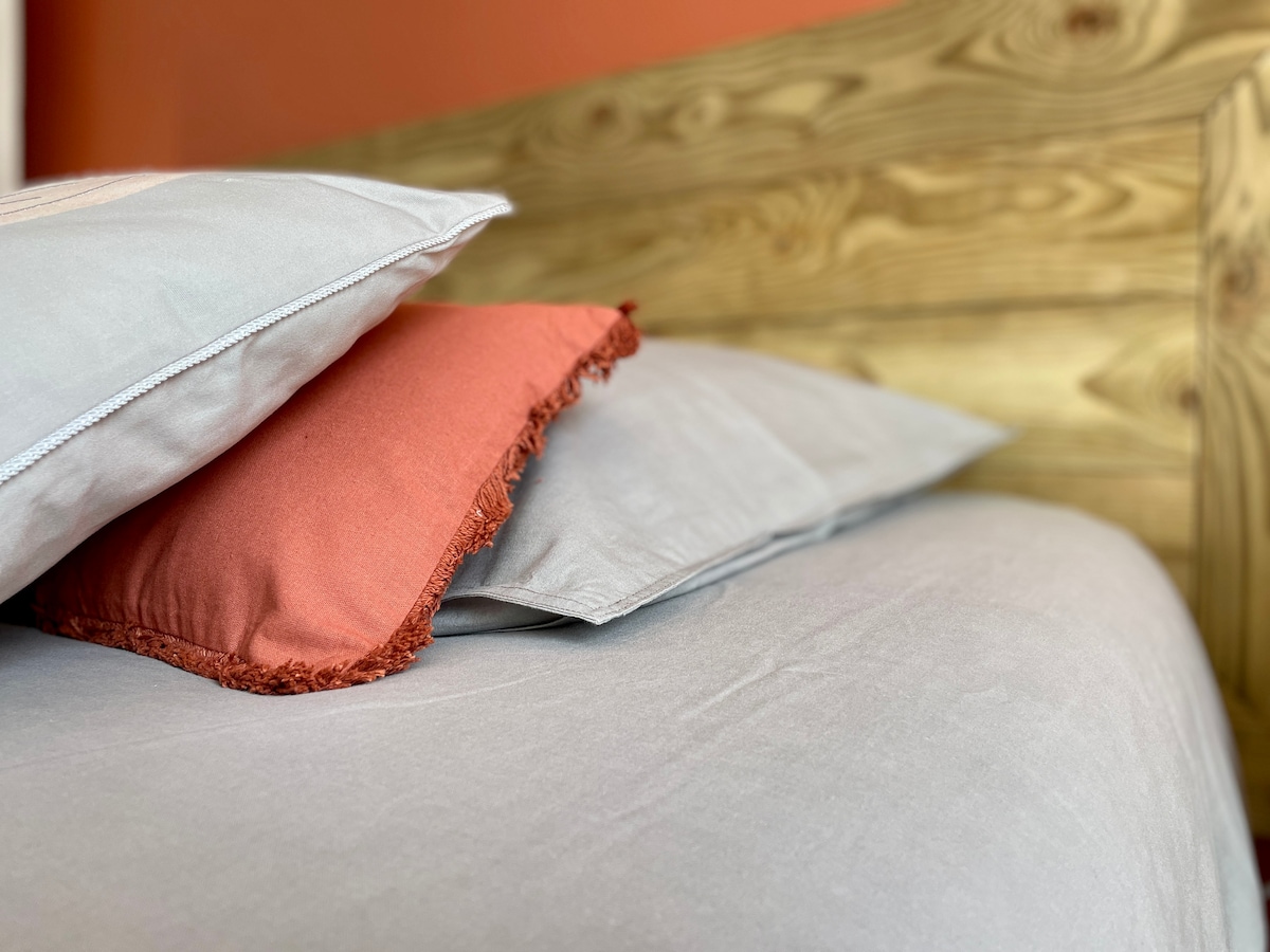 A close-up view of a spacious bed featuring a wooden headboard. Two pillows are arranged on the bed, one is a neutral shade while the other has a rust-colored trim. The bedding appears soft and inviting.