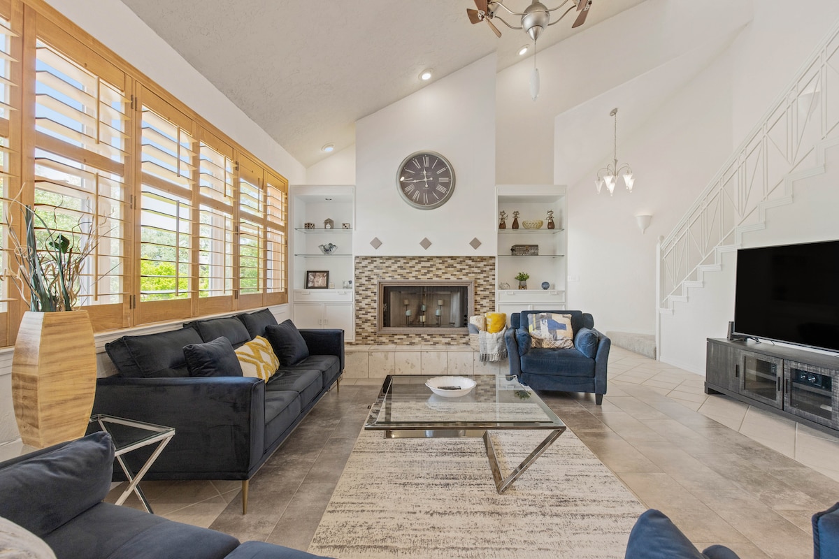 A spacious living area features a large sectional sofa paired with an armchair, positioned around a glass coffee table. Natural light floods in through window shutters, highlighting a decorative fireplace flanked by built-in shelves. A contemporary clock adorns the wall, enhancing the inviting ambiance.