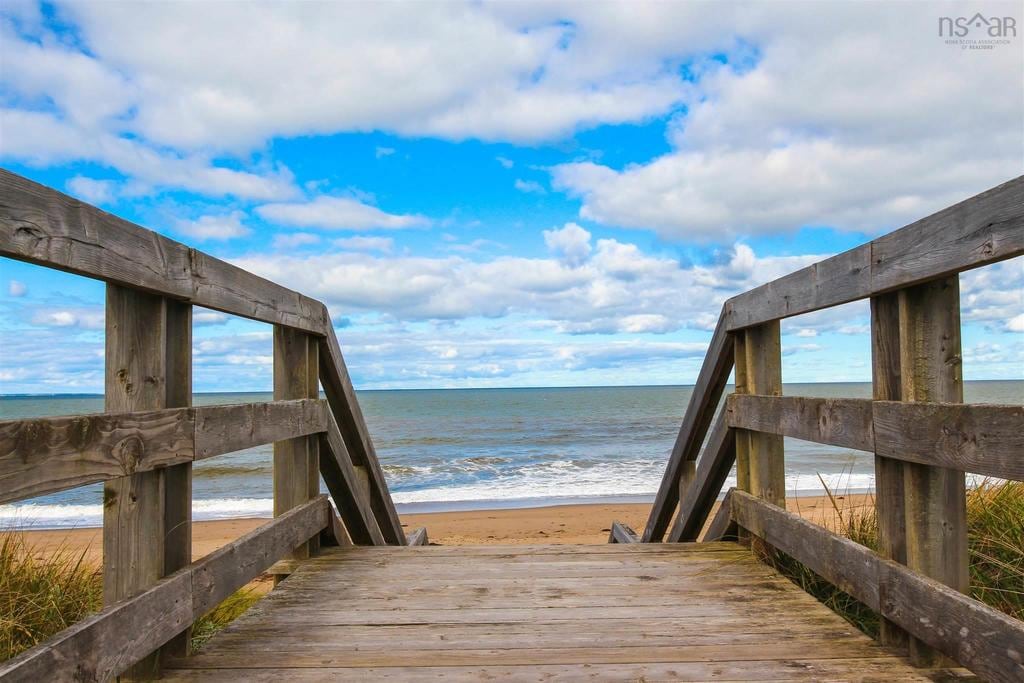A wooden staircase leads to a sandy beach, flanked by wooden railings. The ocean is visible in the distance under a sky filled with scattered clouds. The tranquil scene invites relaxation by the shore.