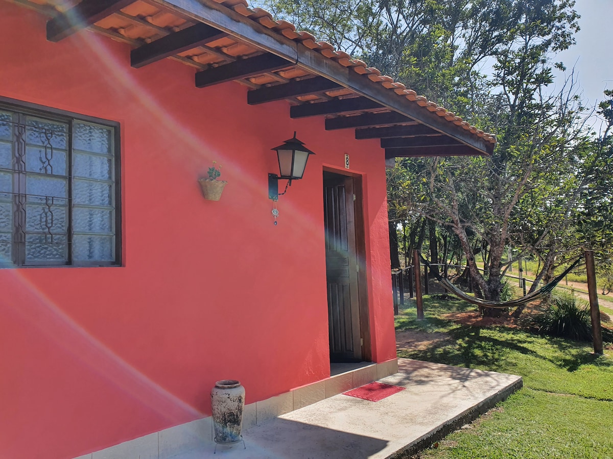 A vibrant red exterior of the chalet is visible, featuring a wooden door and traditional lantern. A small flower pot adorns the wall, while greenery and sunlight create a serene outdoor setting with a hammock in the background.
