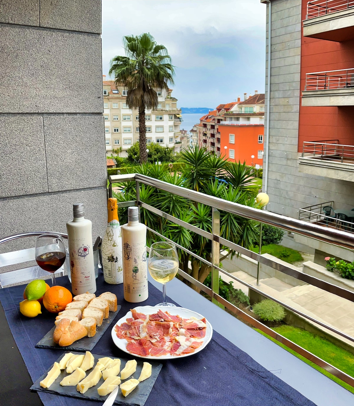 A dining table is set on a balcony, featuring a selection of bread, cheese, and cured meats, accompanied by bottles of wine and glasses. Lush greenery and buildings are visible in the background, with hints of a distant seascape beyond.